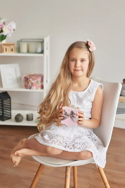 girl in a white dress sits on a chair and holds a gift for mom