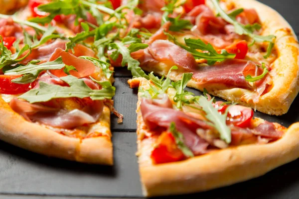 Geleneksel İtalyan pizzasını jamon, roka ve koyu ahşap arka planda vişneli domatesle kapat. Pizza dilimi tabağın yakınına taşınmış. Taze bitkilerle nefis Akdeniz yemekleri.