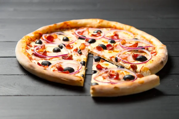 Close up of traditional Italian pizza with ham, sausages, red onion, black olives and tomato sauce on dark wooden background. Slice of pizza moved near the whole dish. Fast food Delicious Still