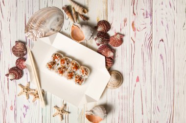 Geleneksel Japon volkanik suşi rulosu yemek çubuklarıyla karton teslimat kutusunda. Ahşap zemin üzerinde deniz kabukları ve yıldızlar. Çiğ somonlu Asya yemeği, uçan balık yumurtası (Tobiko) Unagi sosuyla servis edilir.