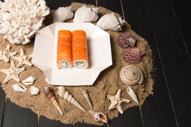 Üzerinde Philadelphia peyniri, salatalık ve çiğ somon filetosu olan Japon suşi rulosu. Okyanus mercanı, deniz kabuğu ve yıldızlı kumun üzerinde Asya yemeği. Siyah tahta arkaplan