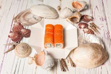 Üzerinde Philadelphia peyniri, salatalık ve çiğ somon filetosu olan Japon suşi rulosu. Okyanus mercanı, deniz kabuğu ve ahşap zemin üzerinde yıldızlar olan paket servis kutusunda Asya yemeği.