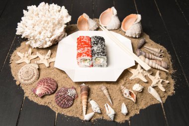 Japon suşi rulosu ve siyah ve kırmızı uçan balık yumurtası (Tobiko) yemek çubuklarıyla paket servis kutusunda. Kızarmış somon, salatalık, krem peynirli Asya yemeği. Kum, deniz kabukları ve yıldızların ahşap arka planı