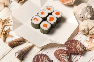 Japon Sake Maki Roll 'un yanına somon balığı ve beyaz karton kutunun üzerinde Norveç yosununa sarılmış pilav. Ahşap zemin üzerinde deniz kabukları ve yıldızlar. Gıda dağıtım kreatice konsept menüsü
