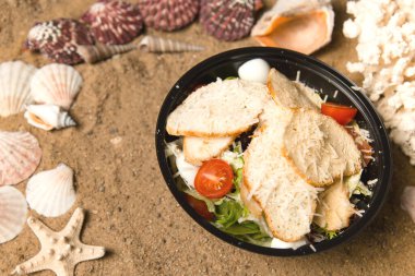 Tavuk fileto, vişneli domates, kızarmış ekmek, bıldırcın yumurtası ve siyah plastik kasede salata karışımı. Yemek paketi ve yemek. Kum arka planında deniz kabukları ve yıldızlar