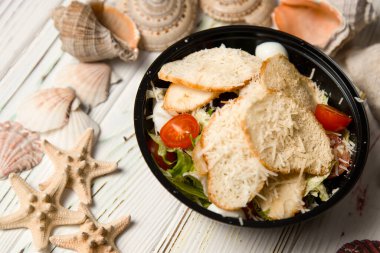 Yakın plan Sezar Salatası tavuk filetosu, kiraz domatesleri, kızarmış ekmek, bıldırcın yumurtası ve siyah plastik kasede salata karışımı. Yemek paketi ve yemek. Tahta arka planda deniz kabukları ve yıldızlar