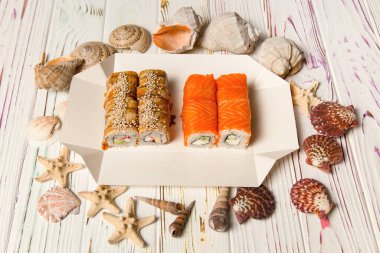İki Japon suşi paketi içinde ahşap arka planda deniz kabukları ve yıldızlarla servis ediliyor. Somon ve yılan balıklı Asya dürümleri. Philadelphia peyniri, salatalık ve avokado..