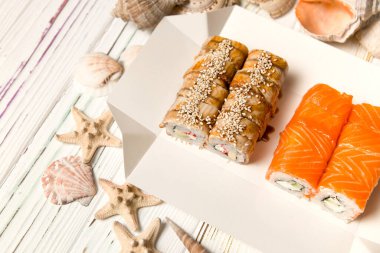 İki Japon suşi paketi içinde ahşap arka planda deniz kabukları ve yıldızlarla servis ediliyor. Somon ve yılan balıklı Asya dürümleri. Philadelphia peyniri, salatalık ve avokado..