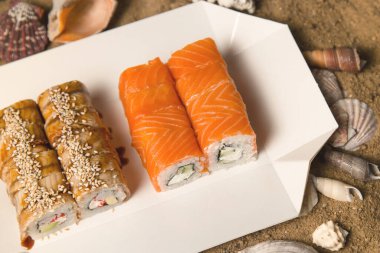 İki Japon suşisi, deniz kabukları ve yıldızlarla kumlu arka planda paket servis kutusunda. Somon ve yılan balıklı Asya dürümleri. Philadelphia peyniri, salatalık ve avokado..