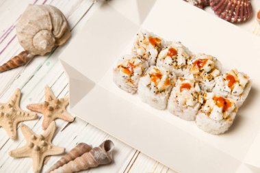 Geleneksel Japon volkanik suşi rulosu beyaz karton teslimat kutusunda. Ahşap zemin üzerinde deniz kabukları ve yıldızlar. Çiğ deniz tarağı, uçan balık yumurtası (Tobiko havyarı) ile Asya yemeği Unagi suce ile servis edilir.