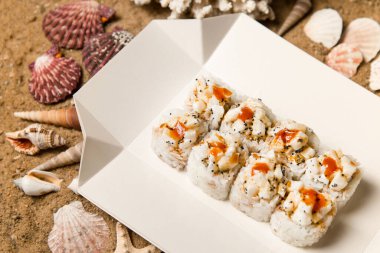Japon volkanik suşi rulosu parçaları beyaz karton teslimat kutusunda. Deniz taraklı Asya yemeği, uçan balık yumurtası (Tobiko havyarı) Unagi sosuyla servis edilir. Mercan, deniz kabukları ve kumdaki yıldızlar..