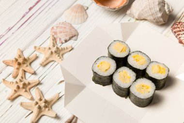 Japon Tamagoyaki Maki Roll, yumurtalı omlet (Tamago) ve Norveç yosununa sarılı pilav beyaz karton kutu üzerinde. Ahşap zemin üzerinde deniz kabukları ve yıldızlar. 