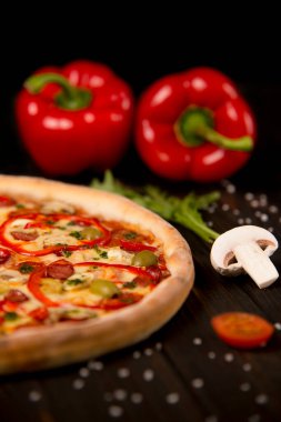 Biberli yarım pizza, yeşil zeytin, sosis, mozzarella peyniri, pepperoni, salam, kırmızı biber, ahşap tahtada iki dolmalık biber. Kesilmemiş pizzaya yaklaş