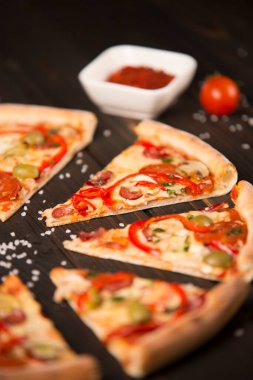 Biberli dilimlenmiş pizzanın yarısı, zeytin, sosis, mozzarella peyniri, pepperoni, salam, biber, ahşap tahtada şampiyonlar, odak noktası deniz tuzu ve arka plandaki malzemeler..