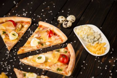 Domatesli pizza, pesto sosu, pastırma, bıldırcın yumurtası, mozzarella ve ahşap zemin üzerinde deniz tuzu ve malzemelerle parmesan peyniri. Pizza parçalarının yanında üç çeşit peynir ve bıldırcın yumurtası.