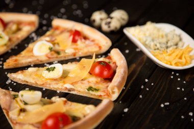 Domatesli pizza, pesto sosu, pastırma, bıldırcın yumurtası, mozzarella ve ahşap zemin üzerinde deniz tuzu ve malzemelerle parmesan peyniri. Pizza parçalarının yanında üç çeşit peynir ve bıldırcın yumurtası.