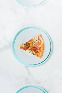 Maydanozlu etli pizza. Domatesli tabak, salam, jambon, şampiyonlar ve Mozzarella peyniri. Pastel turkuaz ve gri servis tabakları üzerine pizza dilimi. beyaz mermer arka planda tablo takımı.