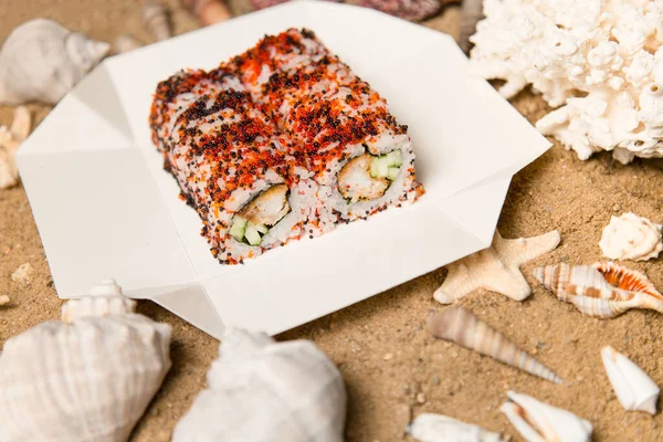 Japon suşi rulosu karton teslimat kutusunda. Kum, okyanus mercanı, deniz kabukları ve arka planda yıldızlar. Çıtır tempura karides, salatalık, pirinç ve renkli uçan balık yumurtası (Tobiko havyarı) ile Asya yemeği.).