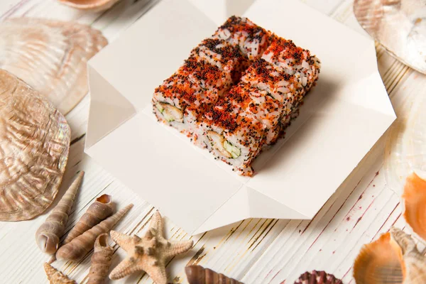 Japon suşi rulosu karton teslimat kutusunda. Tahta arka planda deniz kabukları ve yıldızlar. Çıtır tempura karides, salatalık, pirinç ve renkli uçan balık yumurtası (Tobiko havyarı) ile Asya yemeği.).