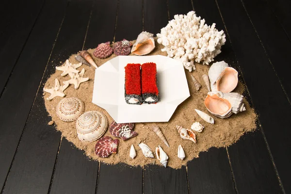 Japanese Sushi Roll in delivery box on wooden board with sand, sea shells and stars. black rice roll with salmon, Philadelphia cream cheese and flying fish roe (Tobiko caviar). Asian menu seafood dish