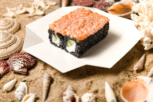 Japon suşi rulosu deniz kabukları ve yıldızlarla kumlu arka planda teslimat kutusunda. Avokado ve Philadelphia krem peynirli siyah pirinç rulosu. Asya menüsünde çiğ somon var.