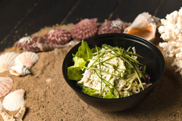 Asian Salad with snow crab meat, cucumber, avocado, flying fish roe (Tobiko caviar) and salad mix in black plastic bowl. Food Delivery package with meal. Sand, sea shells and stars on dark background.
