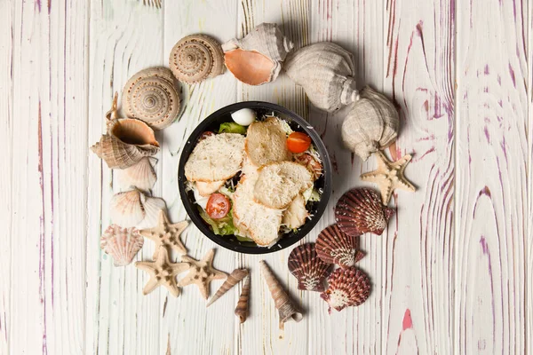 Sezar Salatası 'nın tavuklu fileto, vişneli domates, kızarmış ekmek, bıldırcın yumurtası ve siyah plastik kasede salata karışımı. Yemek paketi ve yemek. Tahta arka planda deniz kabukları ve yıldızlar