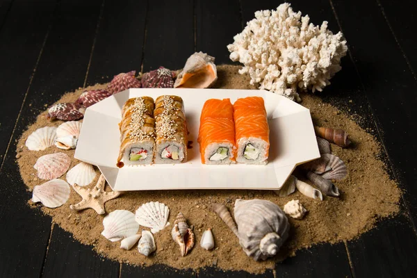 Kum, deniz kabuğu ve yıldızlı ahşap arka planda karton paket servis kutusunda iki Japon suşi rulosu. Somon ve yılan balıklı Asya dürümleri. Philadelphia peyniri, salatalık ve avokado..