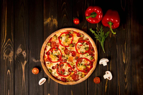 Biberli pizza, yeşil zeytin, füme sosis, mozzarella peyniri, pepperoni, salam, kırmızı biber, ahşap arka planda şampiyonlar. Malzeme ve bitkilerle lezzetli kesilmemiş pizza.