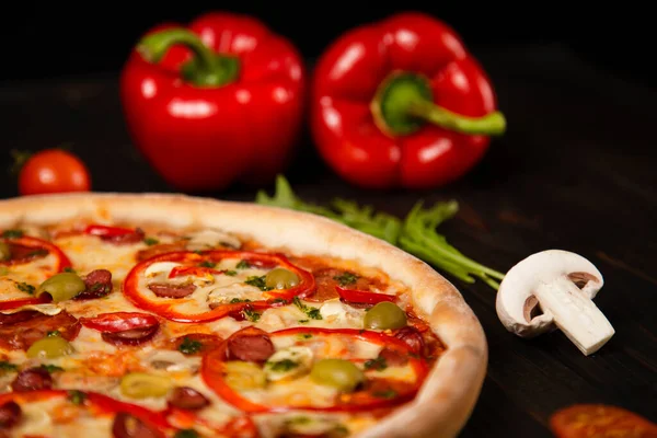 Biberli pizza, yeşil zeytin, füme sosis, mozzarella peyniri, pepperoni, salam, kırmızı biber, arka planda malzemelerle ahşap tahtada şampiyonlar. Kesilmemiş lezzetli pizza.