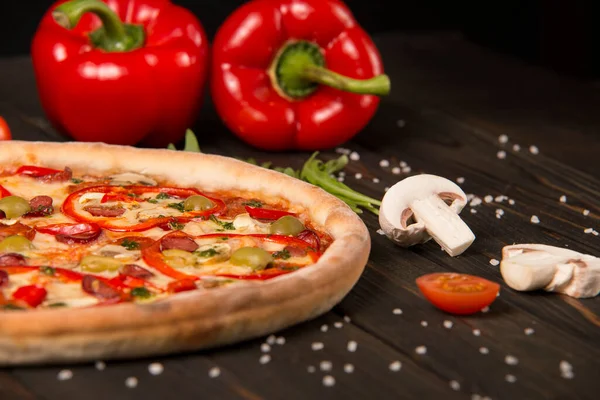 Biberli yarım pizza, yeşil zeytin, sosis, mozzarella peyniri, pepperoni, salam, kırmızı biber, ahşap tahtada iki dolmalık biber. Kesilmemiş pizzaya yaklaş