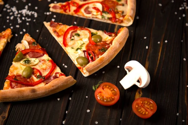 Dilimlenmiş pizzayı dolmalık biber, zeytin, sosis, mozzarella peyniri, pepperoni, salam, biber ve ahşap tahtada deniz tuzu, iki domates ve arka planda şampiyon ile kapatın. Kırsal yiyecekler