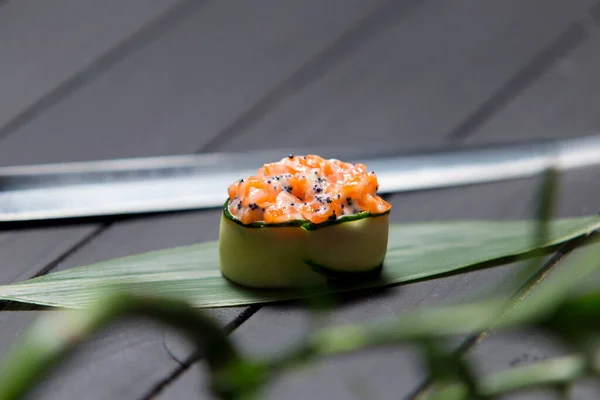 Gunkan Sake Green sushi with salmon and black tobiko caviar served in cucumber on bamboo leaf near Japanese knife. Pan Asian traditional dish on dark wooden board with green plant on foreground.