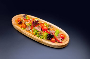 Bitki karışımlı Tayland bifteği salatası ve koyu arkaplanda ahşap tabakta servis edilen kiraz domatesleri. Dana eti parçaları, havuç ve dolmalık biber dilimli taze salata. Restoran menüsü izolasyonu