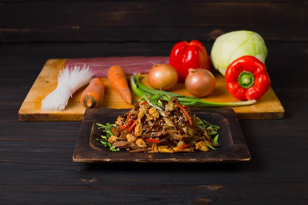 Side view of Harusame Rice noodles with meat served in wooden plate on dark background. Pan Asian dish with carrot and bell pepper slices, sesame seeds, onion scallions. Fresh Vegetables on background