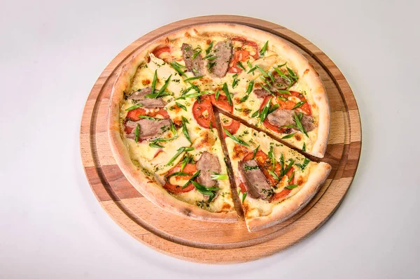 Whole uncut pizza with meat, tomatoes and spring onion scallions  on round wooden cutting board isolated on background. Restaurant fast food menu dish isolation. Slice of meat pizza with melted cheese