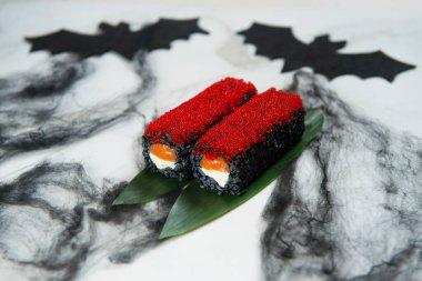 Cadılar Bayramı Tabağı Yemek Sanatı konsepti. Japon Black Rice Sushi Roll bambu yaprakları üzerinde servis edilir, üstünde kırmızı uçan balık yumurtası Tobiko. Siyah örümcek ağı süslemesi ve beyaz mermer arka planda iki yarasa