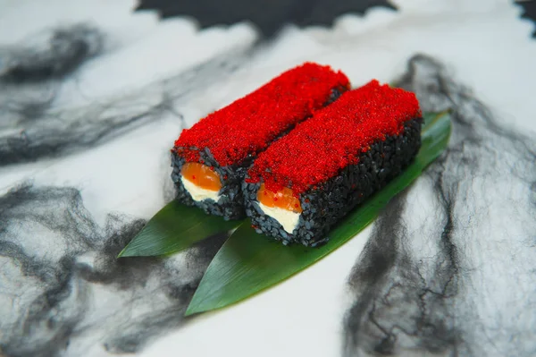 Halloween Holiday Dish Food Art concept. Japanese Black Rice Sushi Roll served on bamboo leaves, red flying fish roe Tobiko on top. Black spider web decoration and bats on white marble background