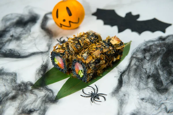 Halloween Holiday Dish Food Art concept. Japanese Black Rice Sushi Roll wrapped in tuna chips Bonita. Black spider and web decoration, Scary painted pumpkin and bat on white marble background