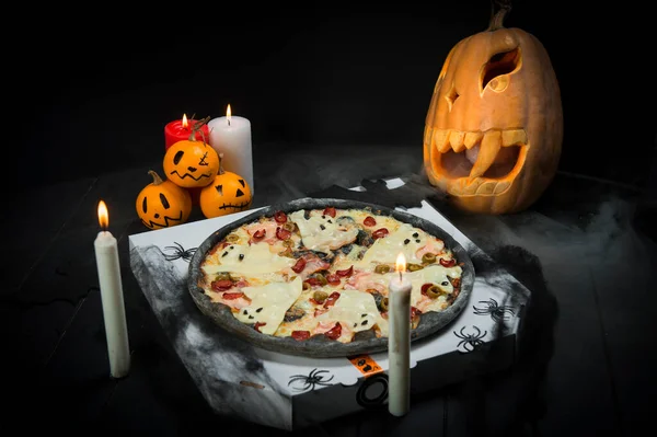 Halloween party decoration. Ghost pizza near spider web, pumpkin jack o lantern And black bats. Candle lights on smoke background. Fast food with cheesy silhouettes served on takeaway pizza box.
