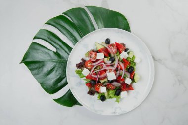 Tabağın altında tropikal canavar yaprağı olan geleneksel Yunan salatasının en üst görüntüsü. Feta peynirli salata, zeytin, marul, taze salatalık, mermer zemin üzerinde kırmızı soğanla servis edilen kiraz domatesleri.