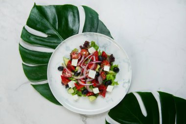 Plakasının altında tropikal canavar yaprakları olan geleneksel Yunan salatası. Feta peynirli salata, siyah zeytin, marul, taze salatalık, mermer arka planda kırmızı soğanlı vişneli domates.