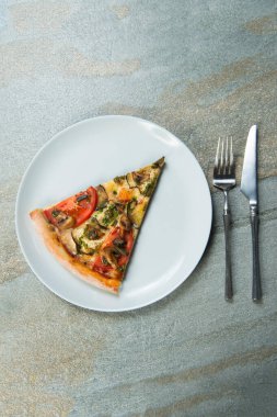 Tabakta sebzeli pizza dilimi. Taş antrasit arka planındaki tabağın yanında çatal ve bıçak var. Domatesli sebze pizzası, şampiyon mantarı, pesto soslu dilimlenmiş patlıcan. Üst görünüm