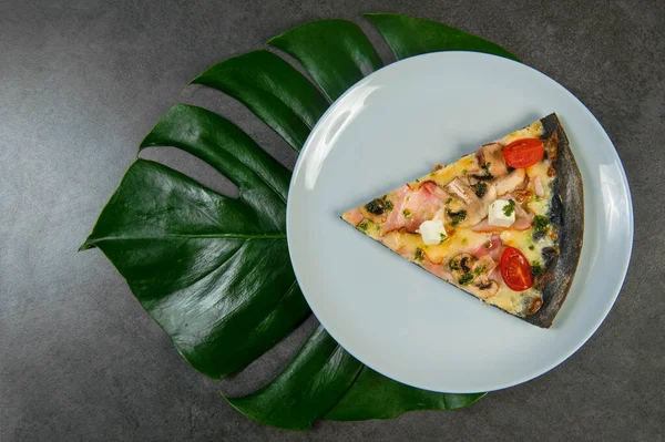 Slice of black dough chicken pizza on plate. Fast food dish on dark stone background. Piece of meat pizza with cherry tomatoes, feta and mozzarella cheese with pesto sauce. Monstera leaf under plate
