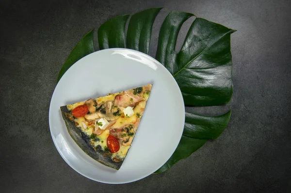 Slice of black dough chicken pizza on plate. Fast food dish on dark stone background. Piece of meat pizza with cherry tomatoes, feta and mozzarella cheese with pesto sauce. Monstera leaf under plate