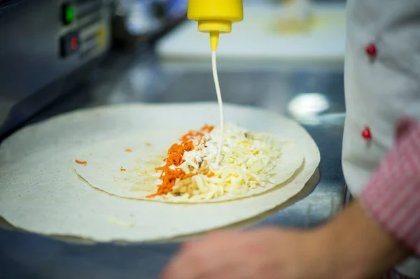 Chef making chicken meat shawarma on professional restaurant kitchen. Lavash bread (pita) with carrot and cheese. Gyros Cooking process. Chef pouring mayonnaise sauce from the bottle. Meat Tortilla