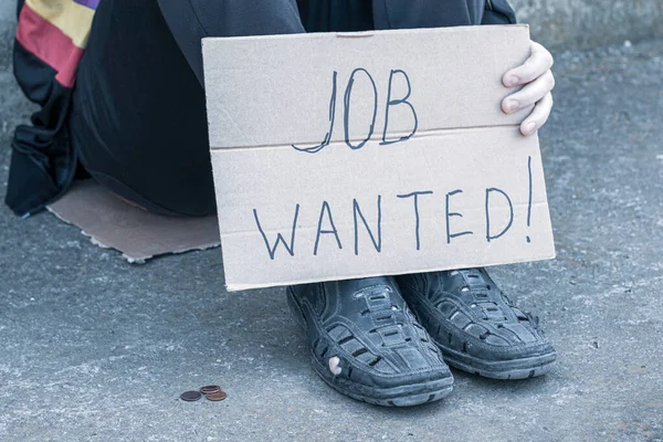 unemployed in old shoes holds in his hand a cardboard tablet with the inscription job wanted on the ground are small coins of alms