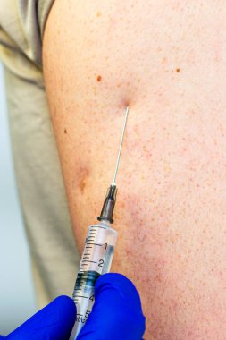 Mavi eldivenli ve beyaz önlüklü doktor hastanın koluna aşı yapar.