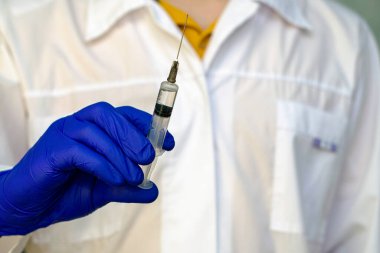 Doktorun elinde beyaz önlüklü bir şırınga. Arka planda bulanık bir doktor figürü.