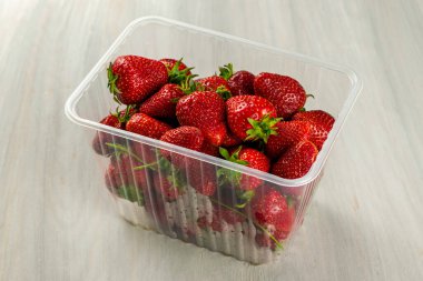 Masanın üzerinde olgun sulu kırmızı çilekler olan şeffaf bir plastik kap var.